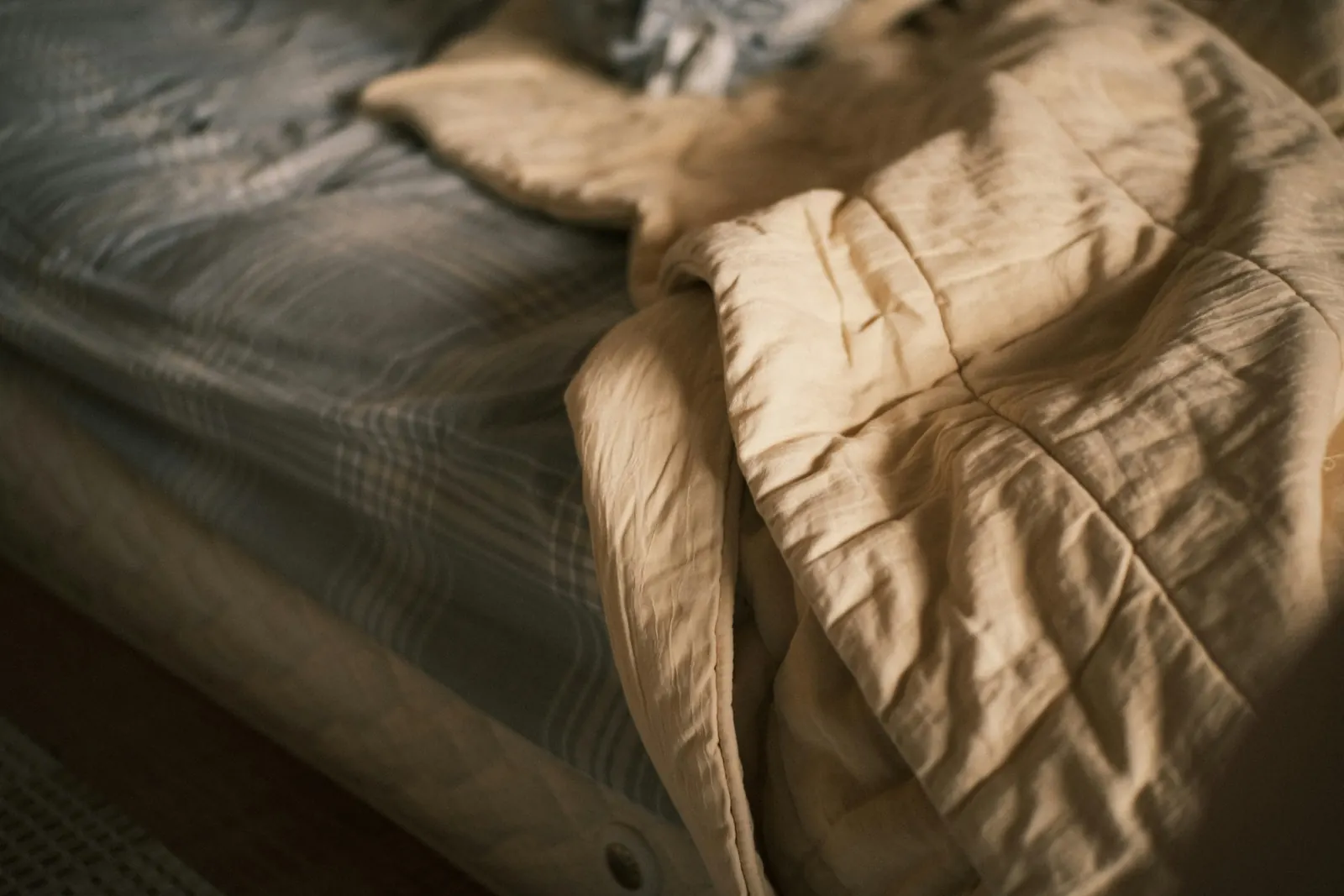 Een onopgemaakt bed met een beige dekbed en gestreepte lakens in zacht ochtendlicht